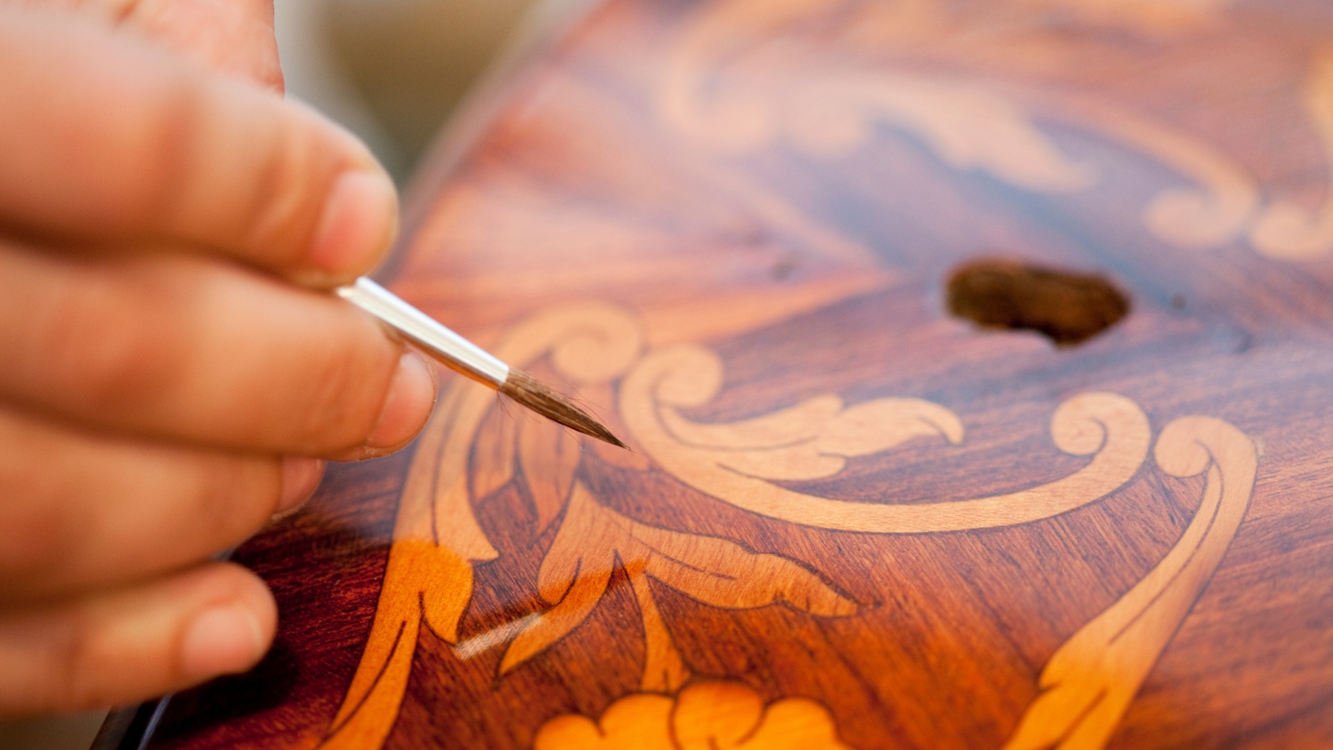 Main d'artisan appliquant du vernis sur une marqueterie de bois aux motifs floraux